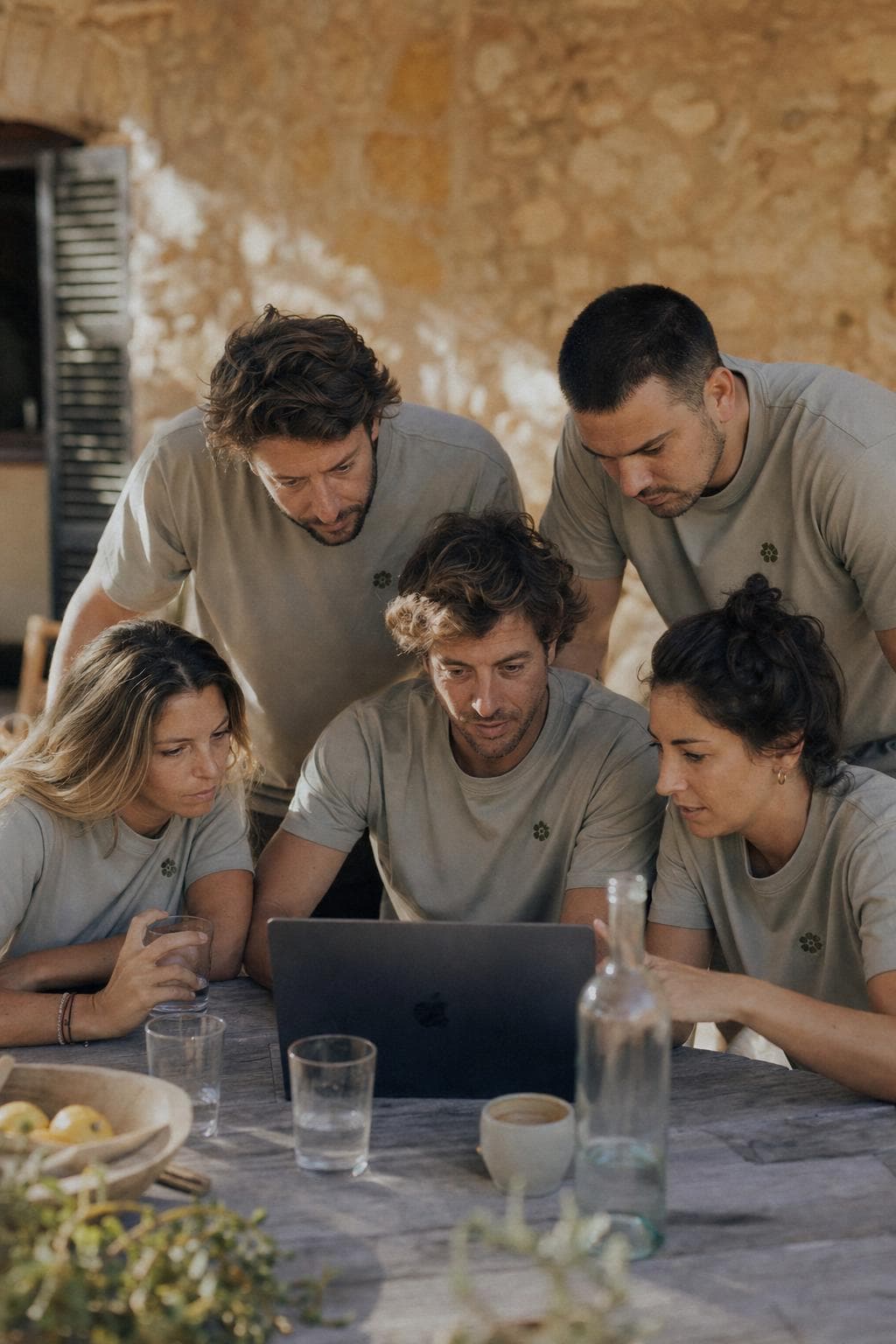 Ein Team mit einheitlichen Firmen-Shirts schaut gemeinsam konzentriert auf einen Laptop auf einer Finca-Terrasse